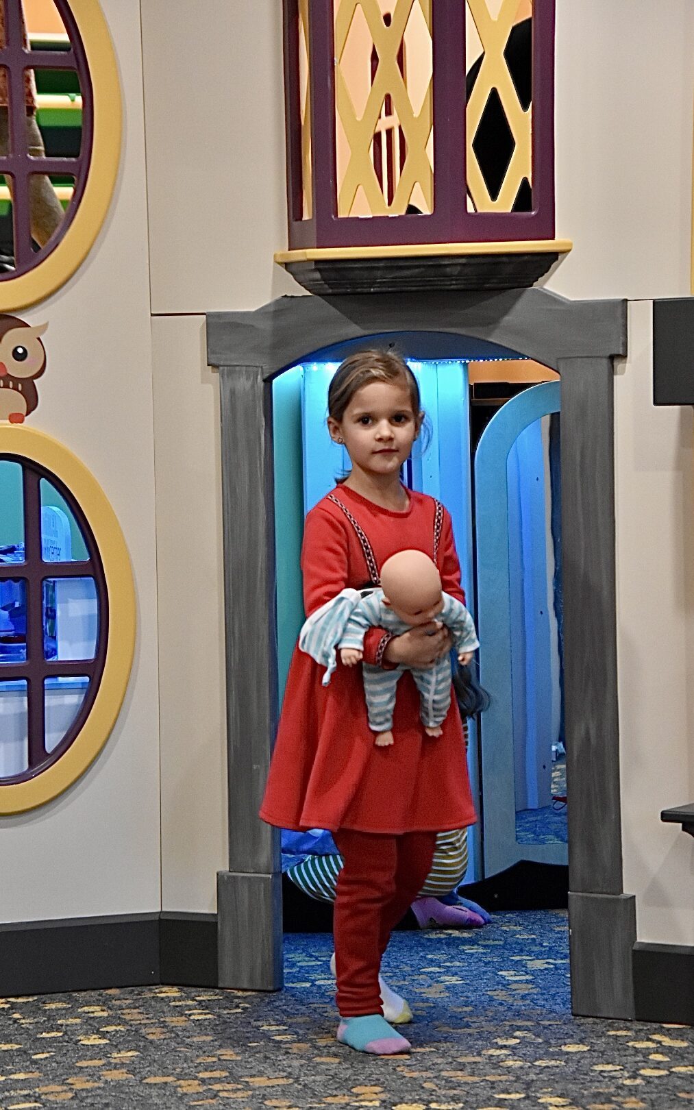 A young guest in a red dress carries her doll through one of the indoor playhouse archways.